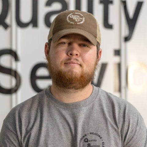 Man in a gray shirt and brown cap standing in front of a blurred sign with partial text.