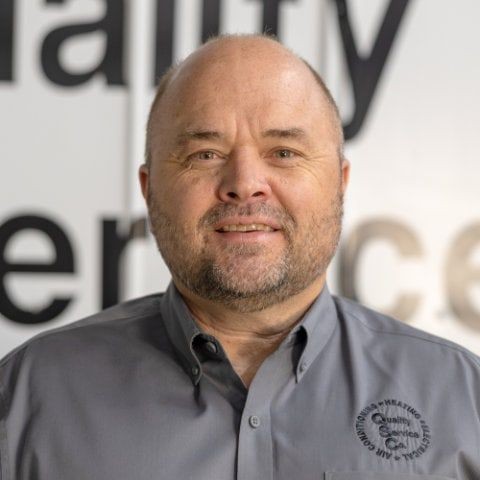 Man with a beard wearing a gray shirt stands in front of a blurred wall with large text.