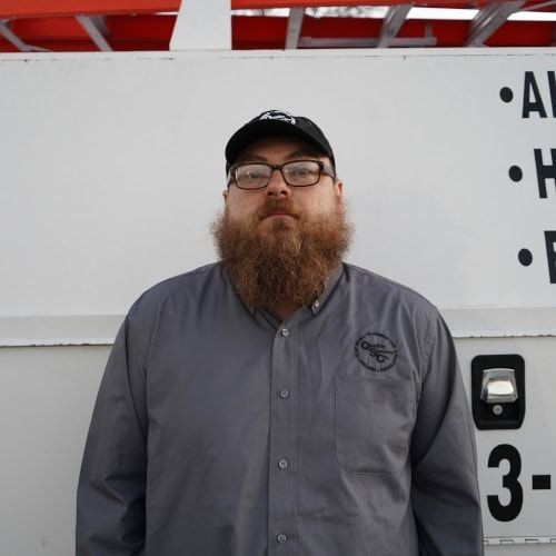 Bearded man in a grey shirt and cap standing against a white background with partial text.