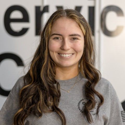 Person smiling in front of a partially visible sign, wearing a gray shirt with curled long hair.