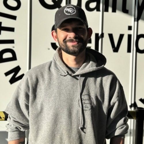 Man wearing a gray hoodie and cap smiling in front of a sign with partial text visible.