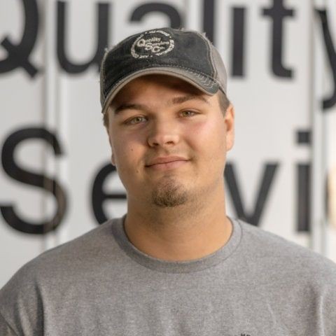 Person wearing a cap and gray shirt smiling in front of a sign with text partially visible.