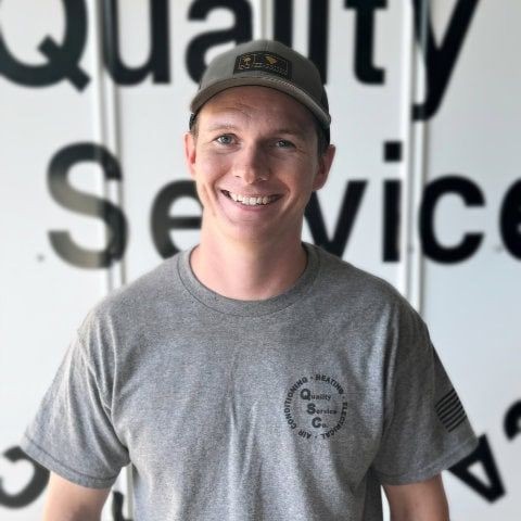 Person wearing a cap and gray t-shirt smiles in front of a sign with partially visible text.