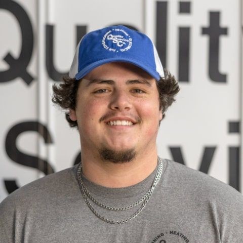 Man wearing a blue cap and gray shirt smiles in front of a wall with partially visible text.