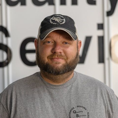 Man wearing a grey shirt and black cap standing in front of a sign with partial text visible.