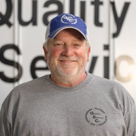 Man wearing a blue cap and gray shirt standing in front of a sign with partially visible letters.
