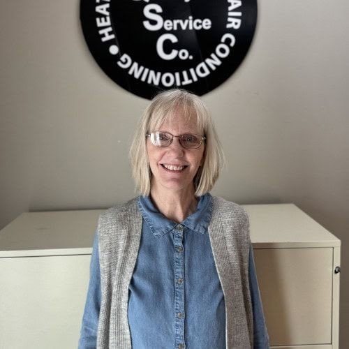 Person with glasses smiling in front of a sign for a heating and air conditioning service company.