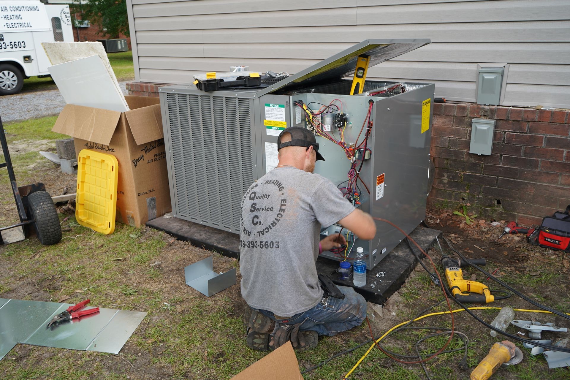 Technician working on an outdoor air conditioning unit beside a house, with tools and wiring visible.