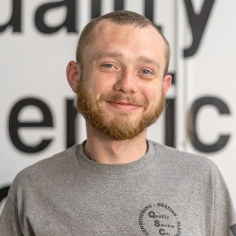Smiling man with a beard wearing a grey shirt, standing indoors against a partially blurred background.