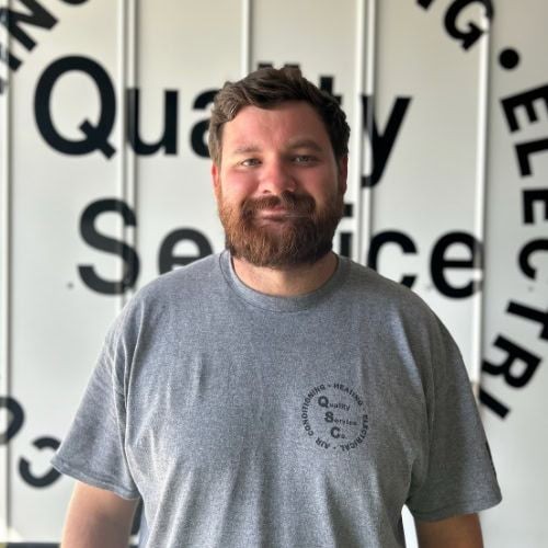 Man with a beard wearing a gray shirt standing in front of a wall with partially visible text.