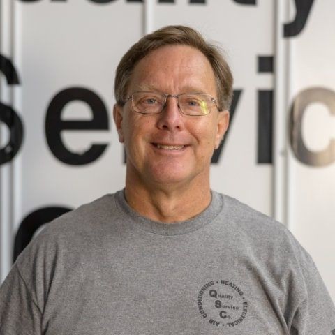 Smiling man wearing glasses and a gray shirt, standing in front of a service-related sign.