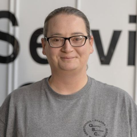 Person wearing glasses and a gray t-shirt standing in front of a wall with partial text visible.