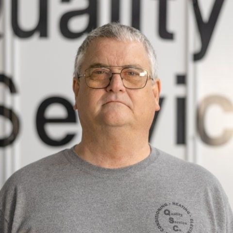 Man with glasses wearing a gray shirt stands in front of a sign with partially visible text.