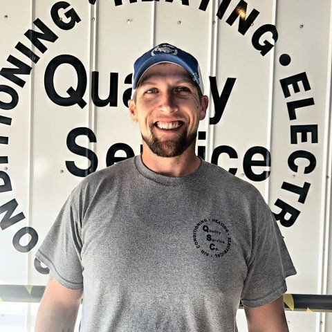 Man wearing a gray shirt and blue cap smiling in front of a service company sign.