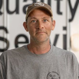 Man wearing a cap and gray shirt standing in front of a sign with partial text visible.
