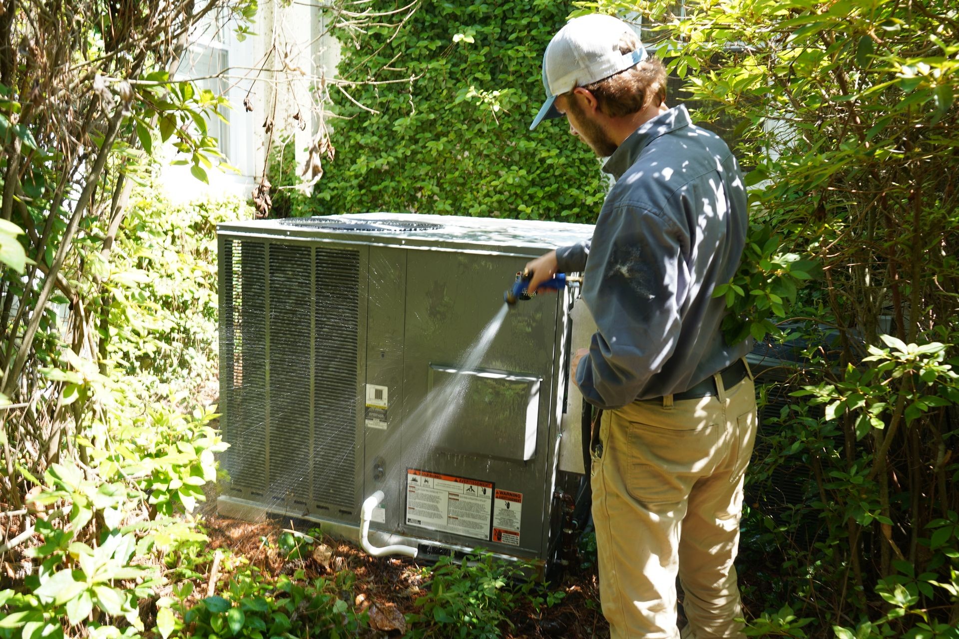 Person cleaning an HVAC unit with a hose in a garden area surrounded by greenery.